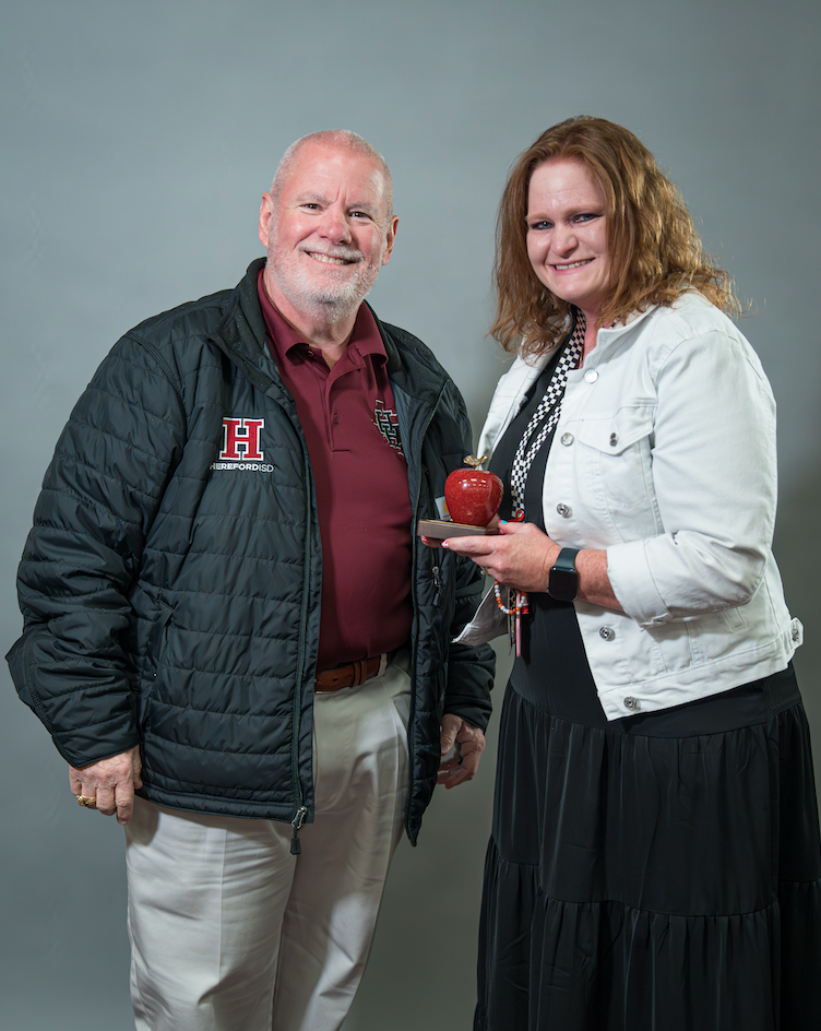 A man and woman stand close, smiling. The man wears a black jacket with an 'H' on the left chest. The woman holds a red apple on a silver plate.