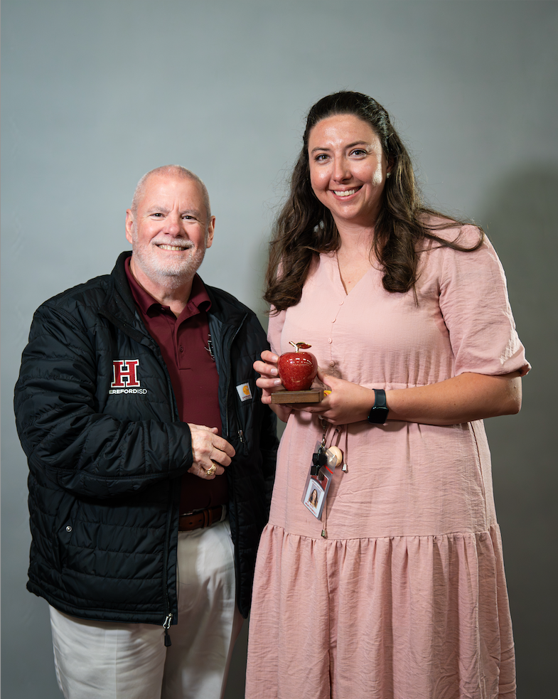 A man and woman stand together. The woman holds a red apple trophy. The man wears a jacket with an H logo.