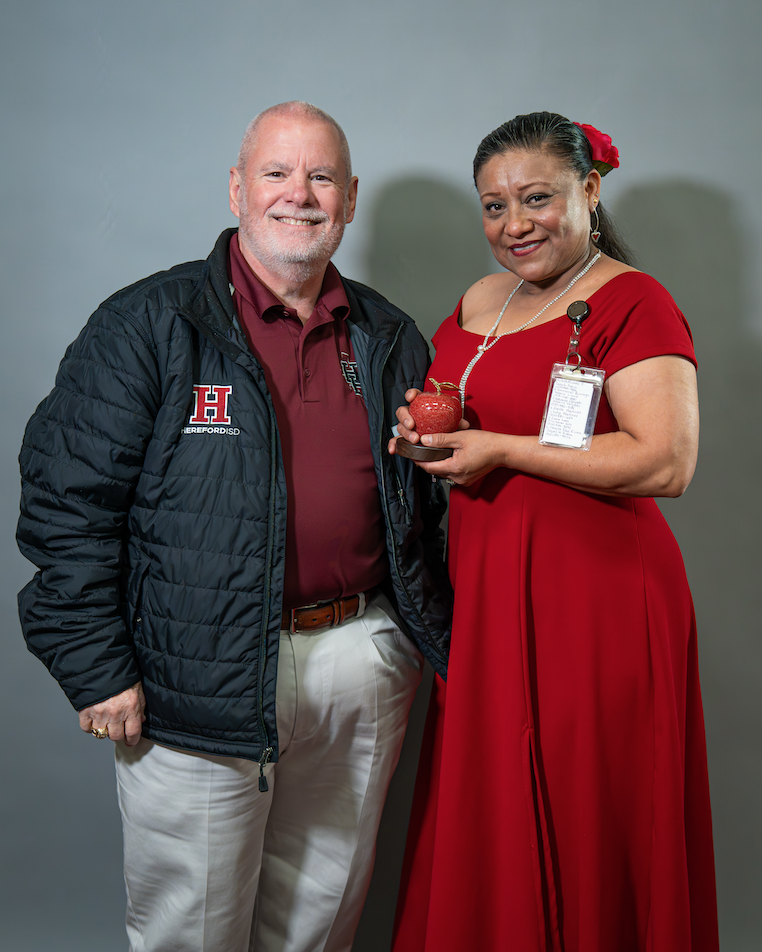 Two people stand together; a man wears a jacket with an "H" logo, and a woman in a red dress holds a small red object.