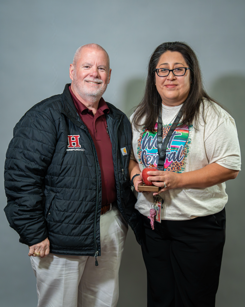 A man and a woman stand side by side with a gray background. The man wears a jacket with an H logo. The woman holds a trophy.