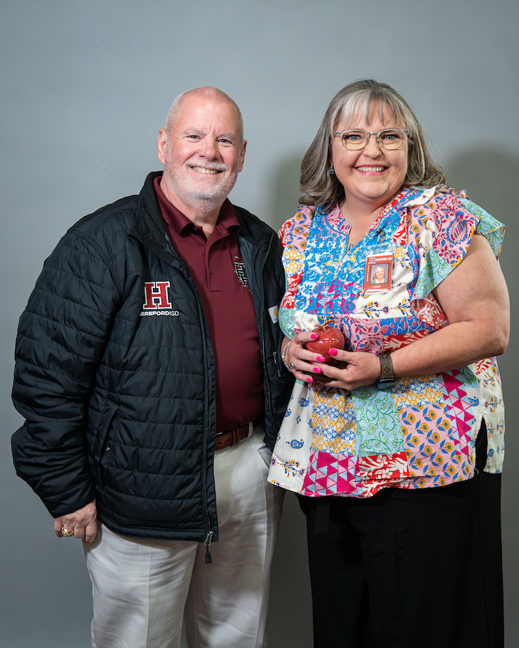 A man and woman standing against a gray background, both smiling. The man wears a black jacket and a red shirt. The woman holds an apple.