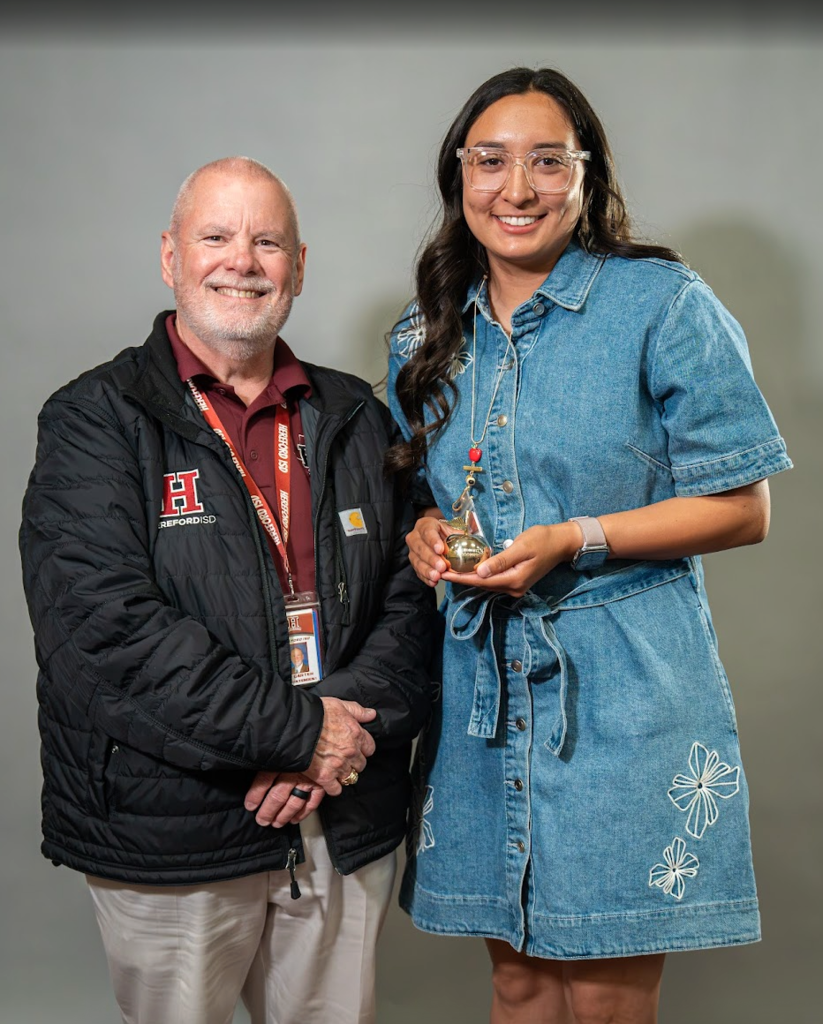 An older man with a name tag smiles and shakes hands with a woman in a denim dress, who holds a small trophy.