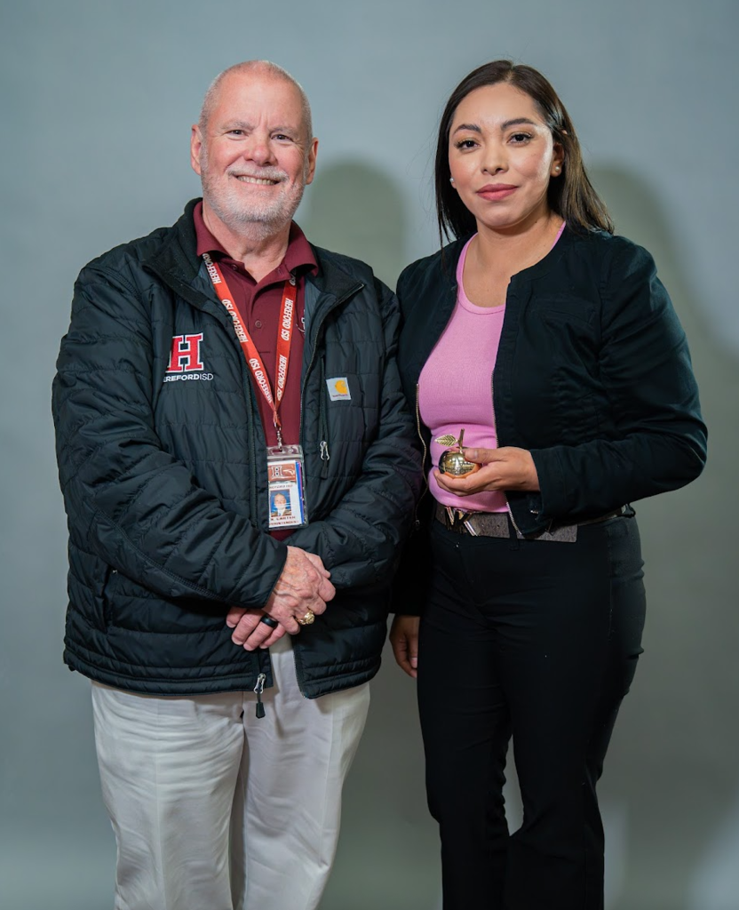 An adult man and woman stand together, smiling. He wears a black jacket with a red ID. She wears a pink shirt and black blazer, holding a trophy.