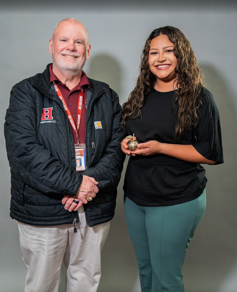 A man and a woman stand next to each other, smiling. The man wears a black jacket, and the woman holds a trophy.