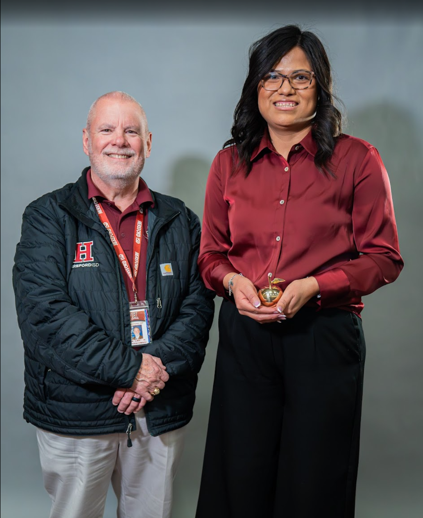 Two people, one with glasses and a red shirt, the other in a black jacket with a badge, standing in front of a gray background.