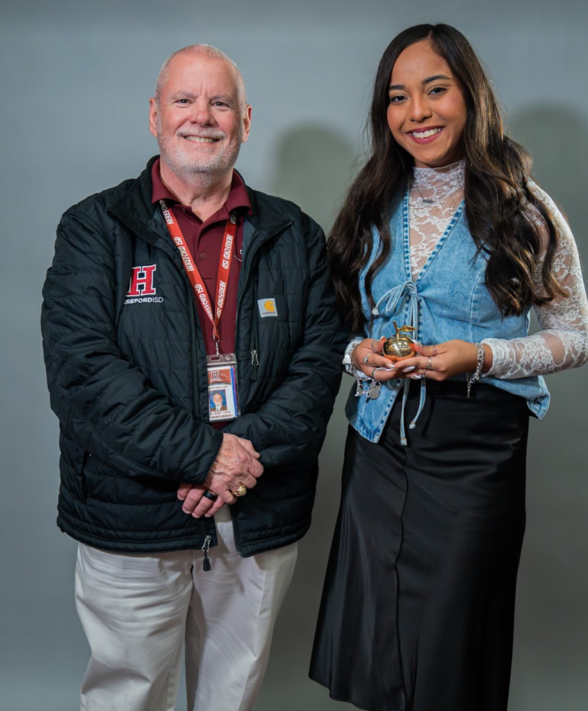 A man and a woman stand together, smiling. The man wears a black jacket and white pants, and the woman a denim top and black skirt.