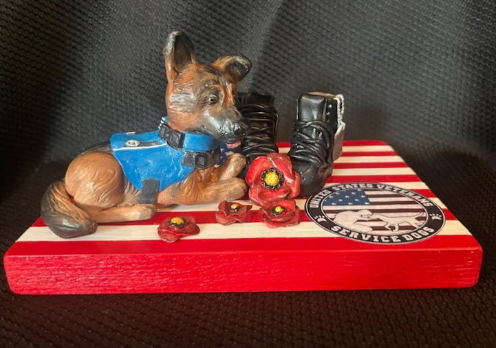 A figurine of a dog in uniform rests on a red and white box. It is surrounded by boots, a collar, and artificial red flowers.