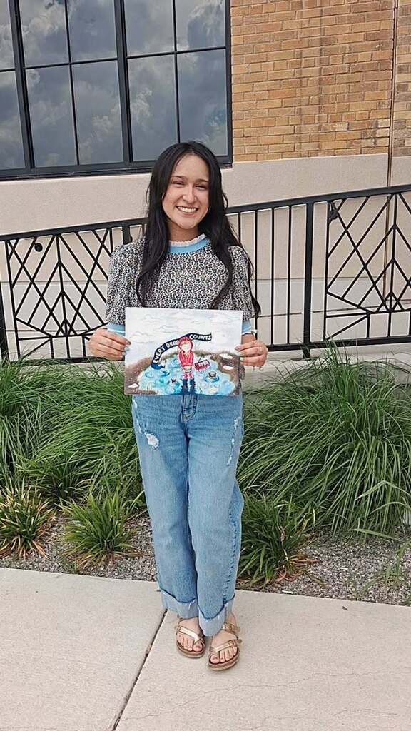 A person with a smile holds a drawing in both hands in front of a building with a brick wall.