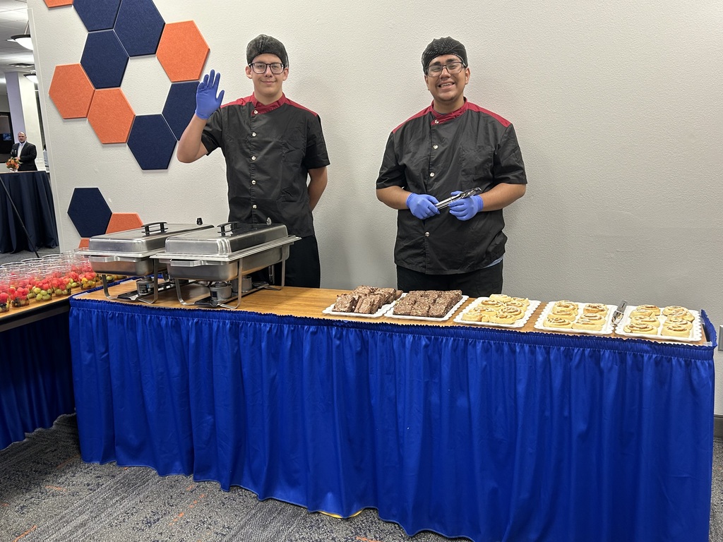 Two people in black uniforms and gloves stand behind a table with desserts on display. Behind them, a colorful wall adds to the event's ambiance.