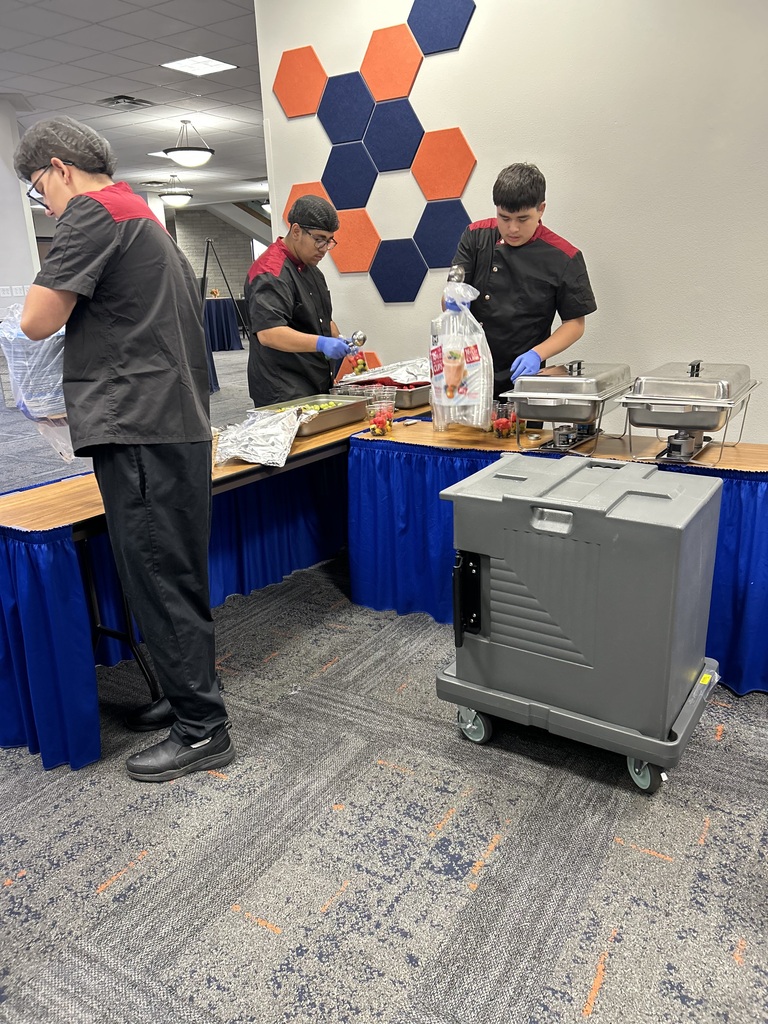 Three individuals stand at a table with blue tablecloths, preparing food. A gray cart is nearby. The wall has hexagonal orange and blue designs.