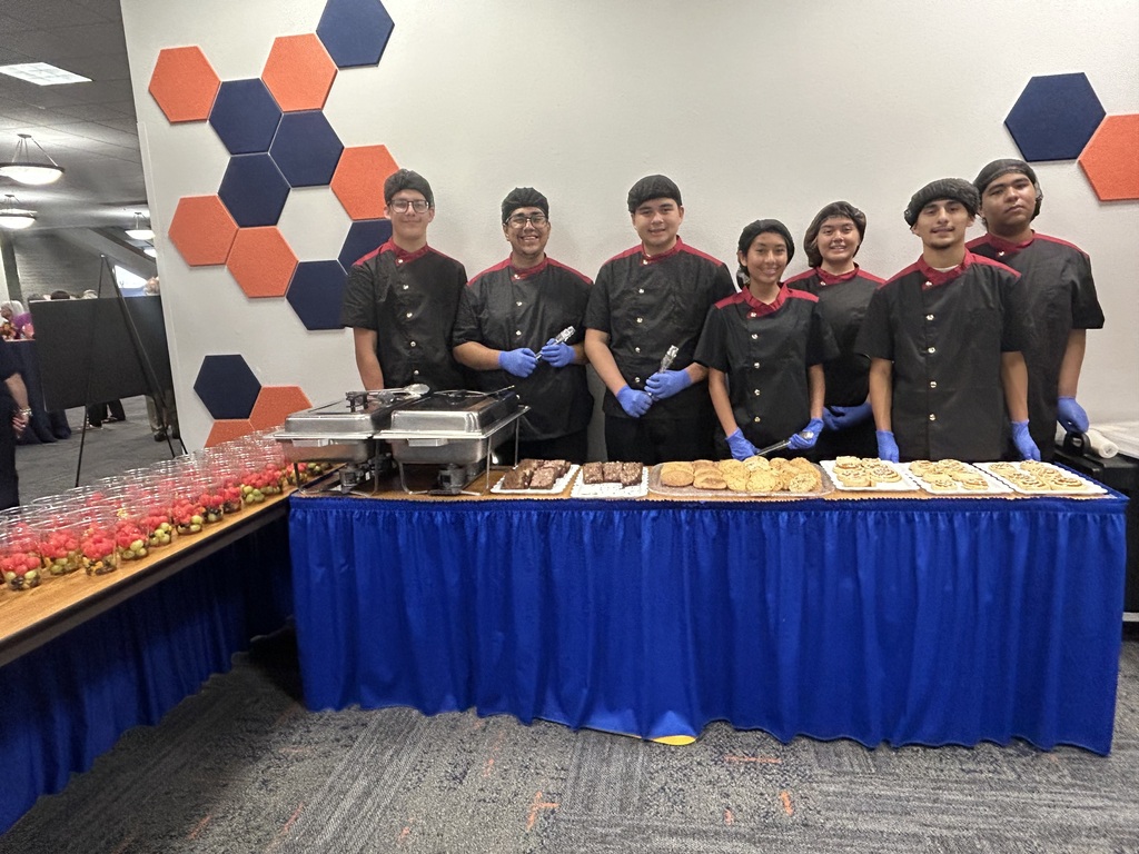 A group of people in matching uniforms, gloves, and hats stand behind a table with a blue cover.
