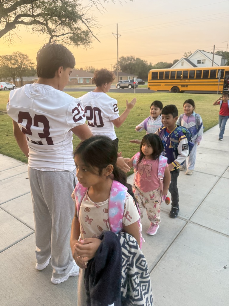 Several children with backpacks walk on the sidewalk as a man stands and talks to them. Behind them, a bus is parked.