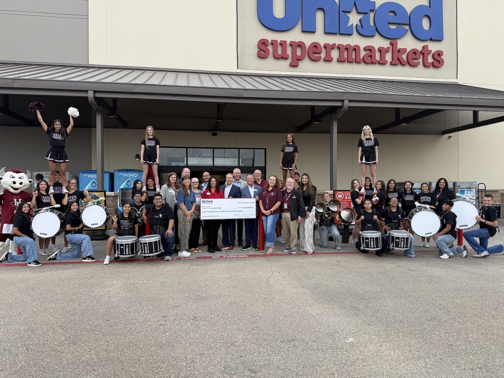 Group in front of a United Supermarkets store. People stand and sit, holding a large check. Three cheerleaders balance atop a stack of drums.