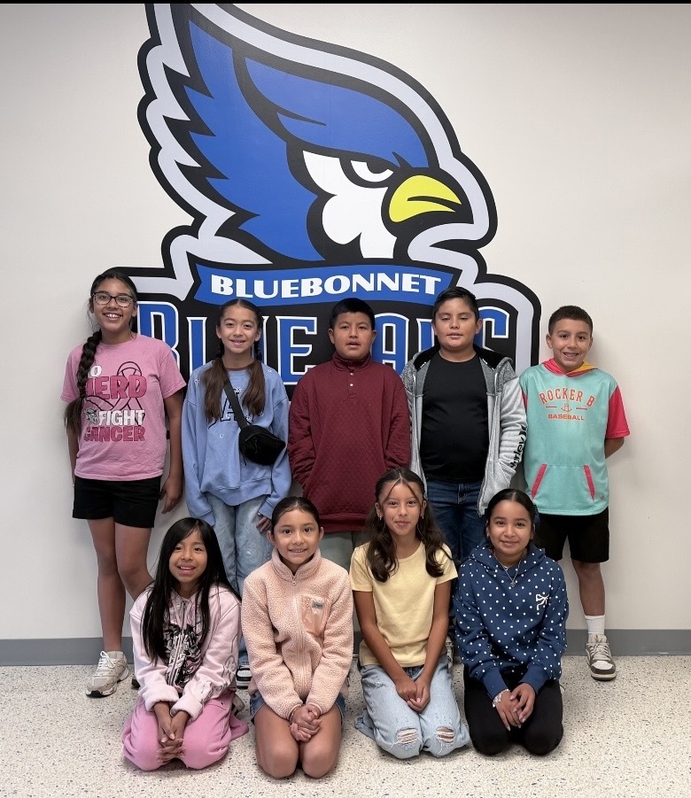 A group of children in various clothes pose in front of a wall with a Bluebonnet school logo.