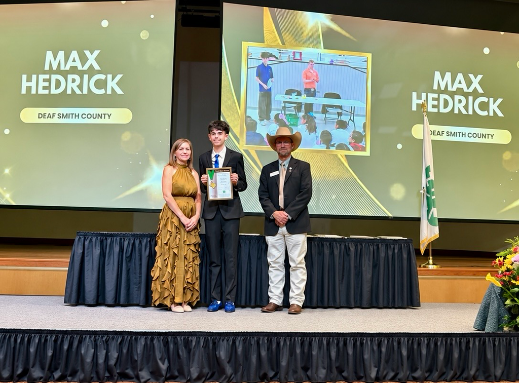 Three people on stage in front of screens, holding certificates and smiling. A man wears a cowboy hat.