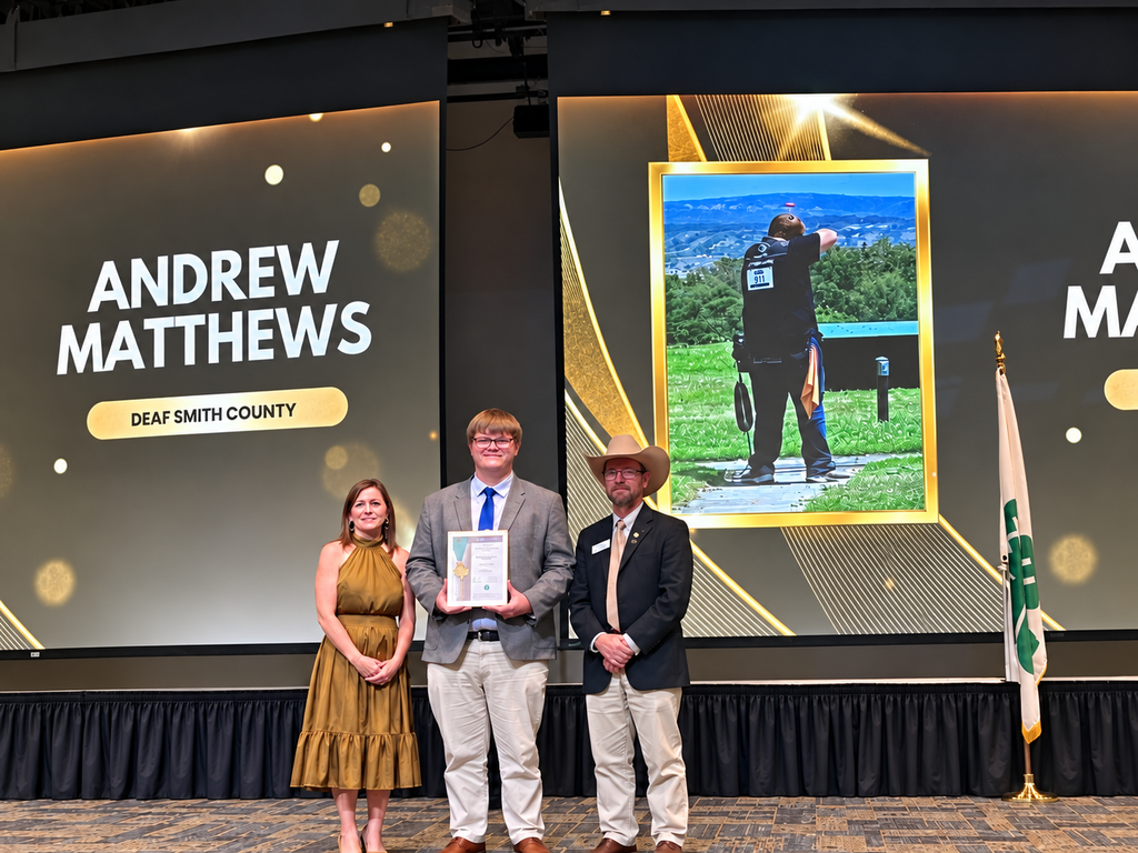 Three people stand on stage; a man holds an award, a woman beside him, and a man in a hat. Behind them, a screen displays a photo of a person and the name "ANDREW MATTHEWS."