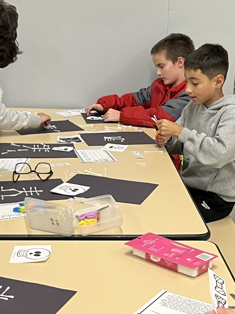 Three boys seated at a table with schoolwork. One holds scissors, another has a book.