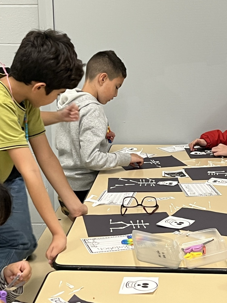 Four students working on a table with papers and colored markers. One is holding scissors.