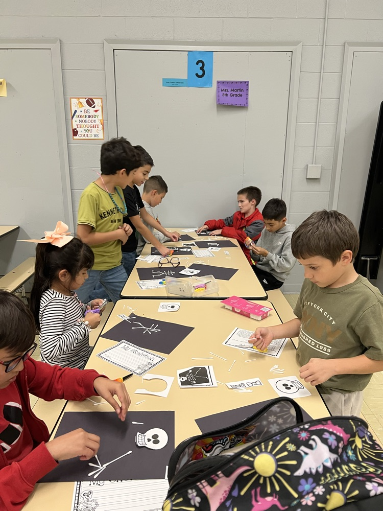 A group of children sit at tables with paper, scissors, and other art supplies.