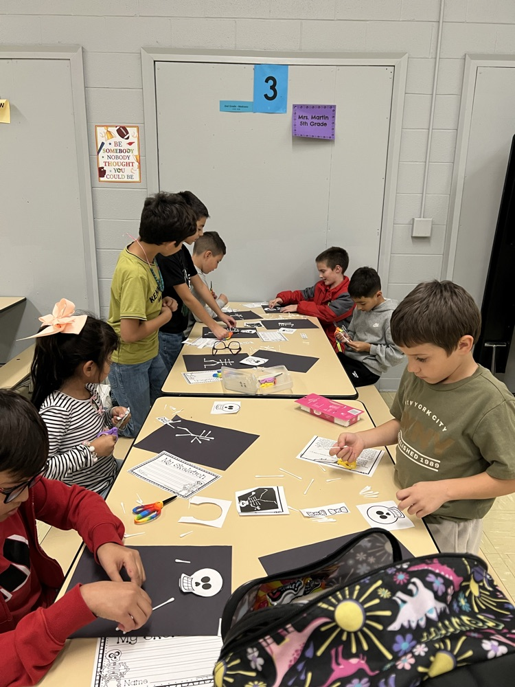 Kids seated at tables with paper, scissors, and crafts. One child is wearing glasses.