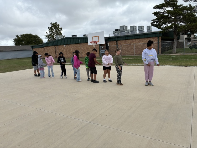 Several students stand in line on a basketball court, facing the basketball hoop, with a building and greenery in the background.