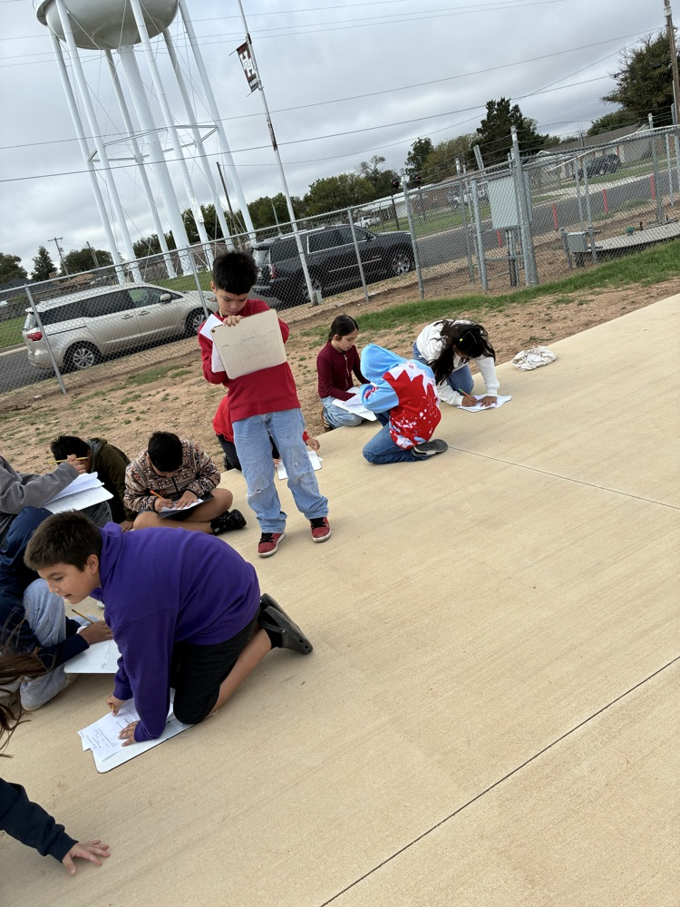 Students outside using clipboards and pencils to write, with a water tower and fence in the background.