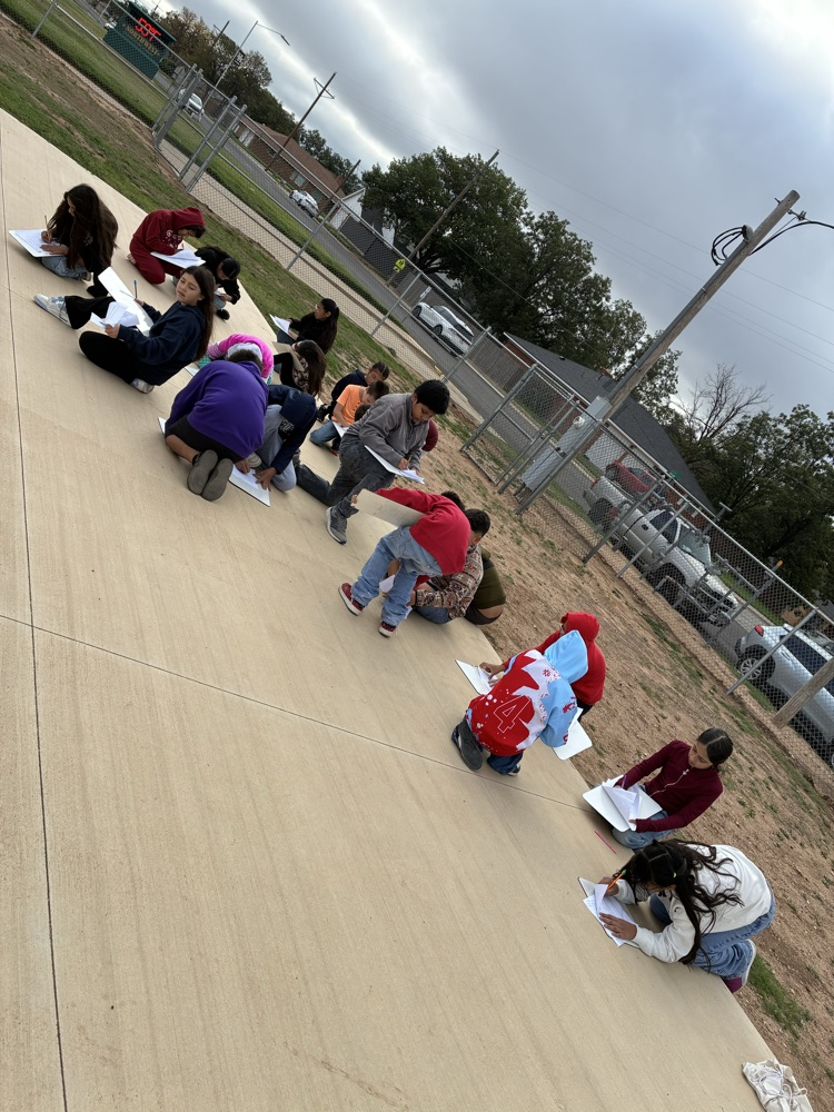 Students sit in a row on a sidewalk, some holding papers, and others looking at papers.