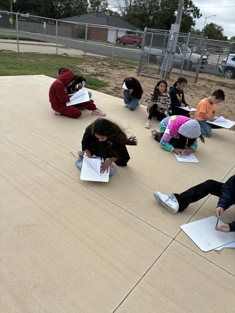 Several students sit on pavement outdoors, writing on paper. Behind them is a fence and parked vehicles.