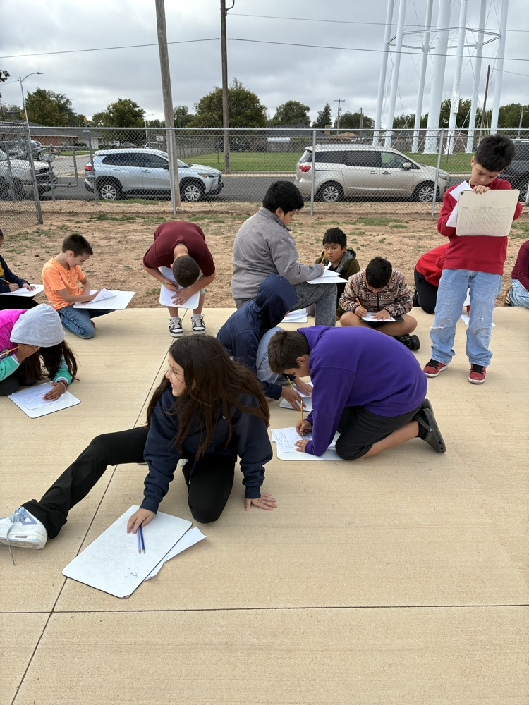 A group of students outdoors, sitting or kneeling on the pavement, holding papers and writing.