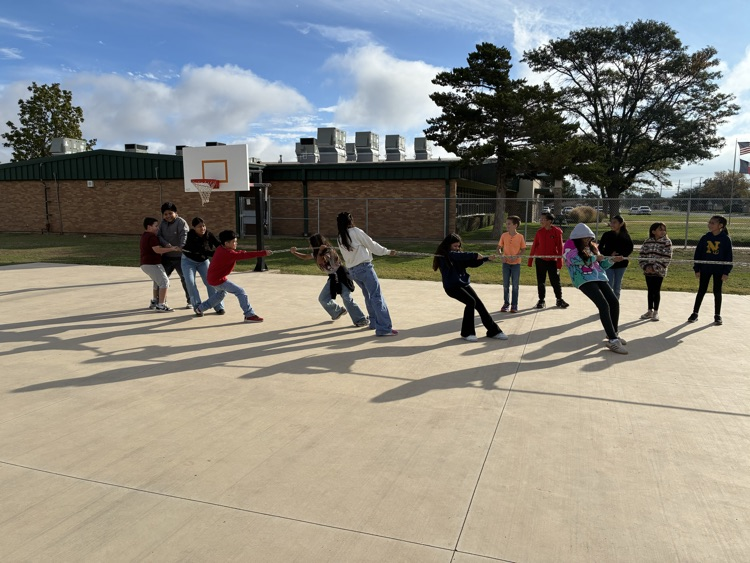 A group of students playing a game on a paved outdoor area with a basketball court.