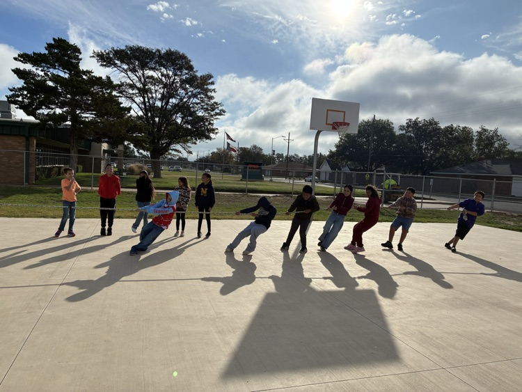 Students are playing a game on a basketball court. Some are bending forward. Trees and a building are in the background.