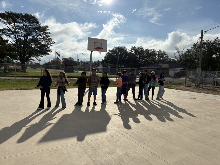 A group of students stand in a line in an outdoor basketball court with a fence.