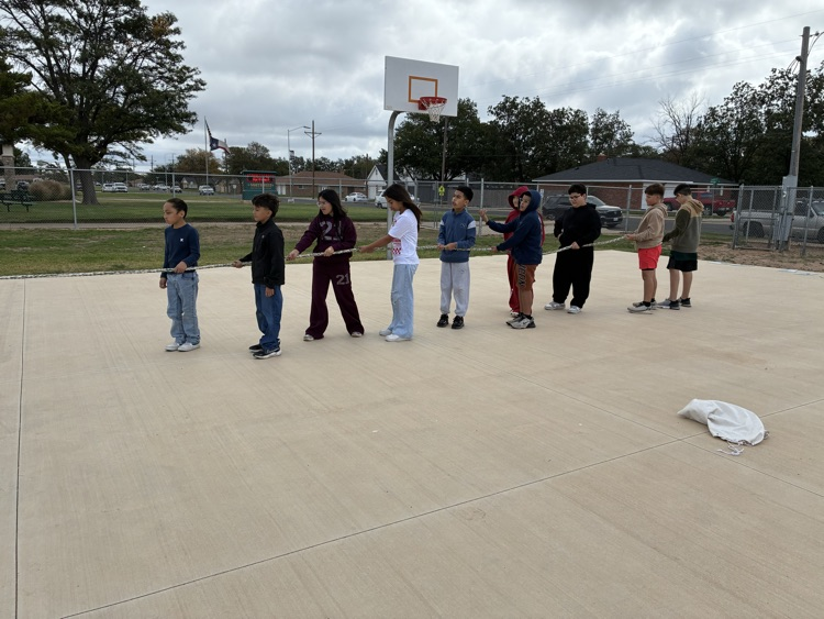 Several children standing in line on a basketball court, holding onto a long rope.