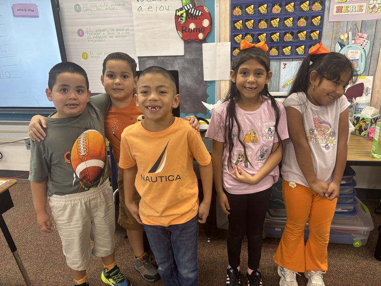 Five children in a classroom. Three boys wearing shirts, one with a graphic, the other two with sleeves rolled up.