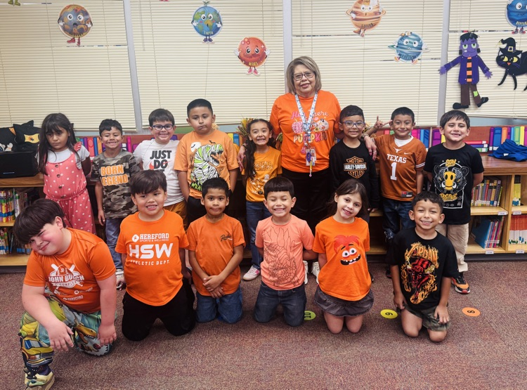 Group of children in orange T-shirts with adult standing in front of a bookshelf.