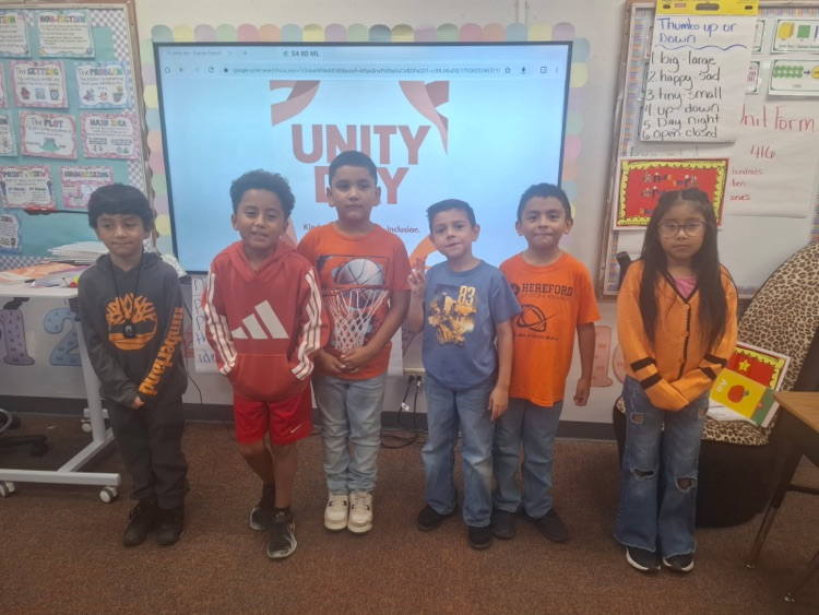 Six children stand in front of a classroom screen showing the words "UNITY BY". Behind them, there are bulletin boards and a small table.