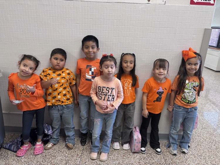 Nine children in orange shirts and jeans standing together in a hallway. Some wear sneakers, others sandals. Some have bags.
