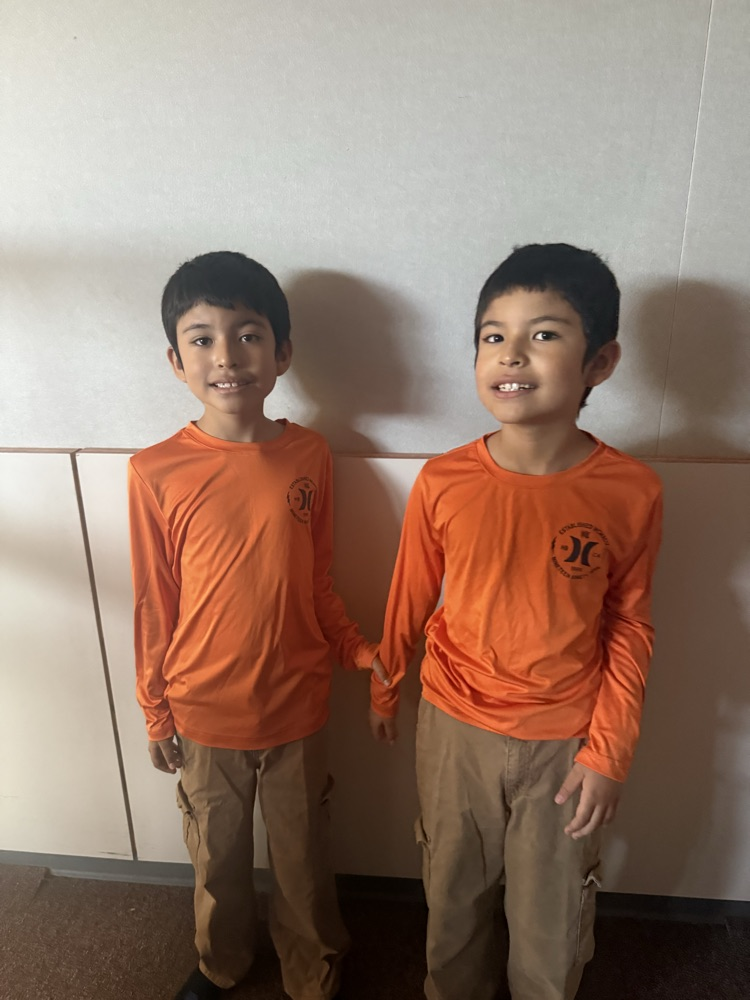 Two young boys in orange long-sleeve shirts and brown pants stand together in front of a white wall.