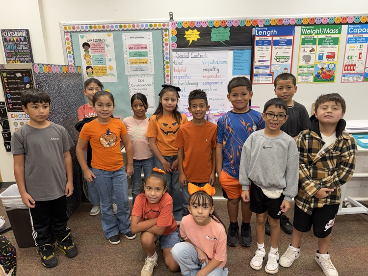 Children in a classroom posing for a group picture. Some wearing orange shirts, glasses, sneakers.