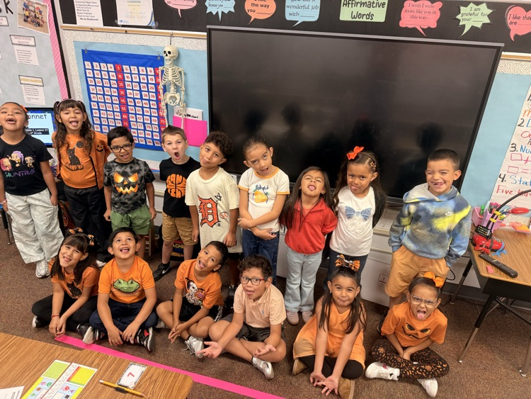 Students posing for a photo in a classroom, smiling, with desks and a large screen behind.