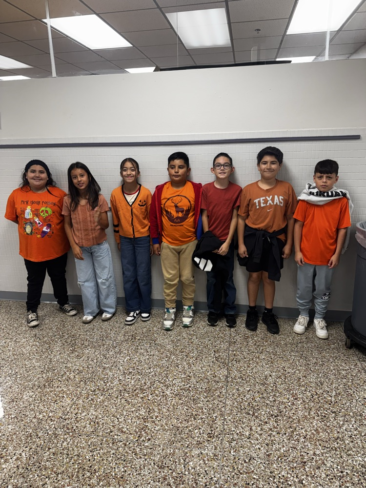 Seven children in orange shirts, one wearing glasses, stand in a line in a white-walled room.