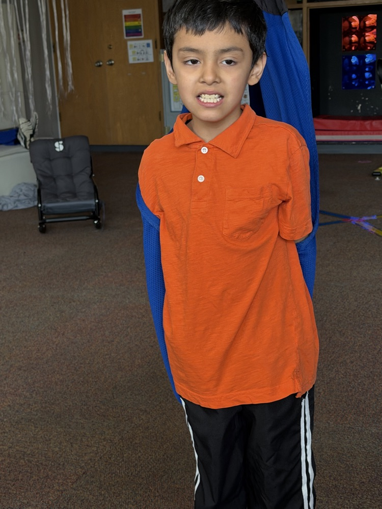Boy wearing orange polo shirt and black pants standing with blue strap over shoulder.