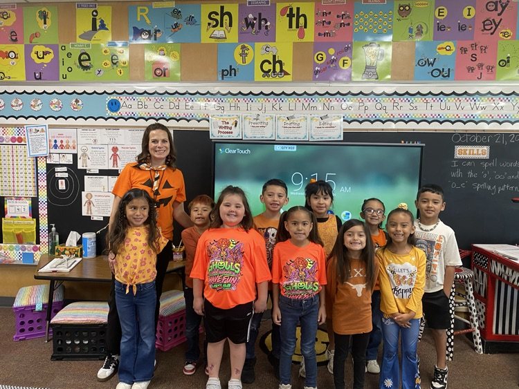 Classroom with students in orange shirts posing with a teacher for a photo in front of a board.