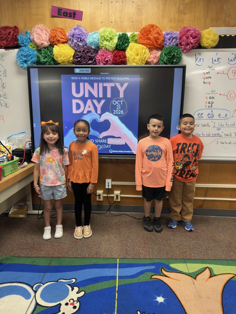 Four children standing in front of a screen with the words "UNITY DAY" and colorful paper flowers.