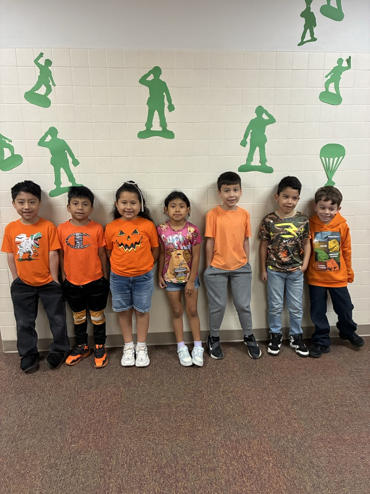 Seven children in orange shirts and sneakers standing in front of a wall with green cutouts.