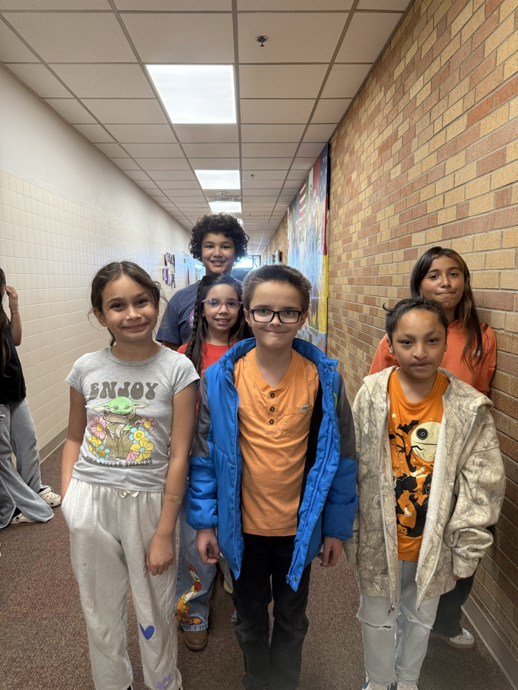 Six children stand in a hallway, smiling, between brick walls. Two children wear glasses, and one wears a jacket.