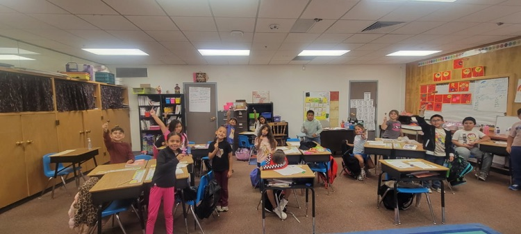 Students stand in a classroom with desks, a teacher, books, and posters on the walls.