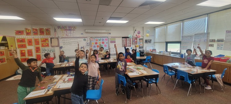 Classroom with students raising hands, desks, tables, chairs, bulletin boards, and a window.