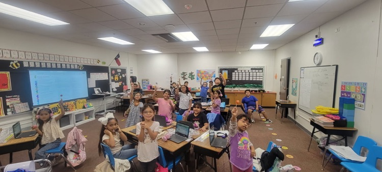Classroom with desks, chairs, and students; some seated, others standing. Laptops on tables, whiteboard nearby.