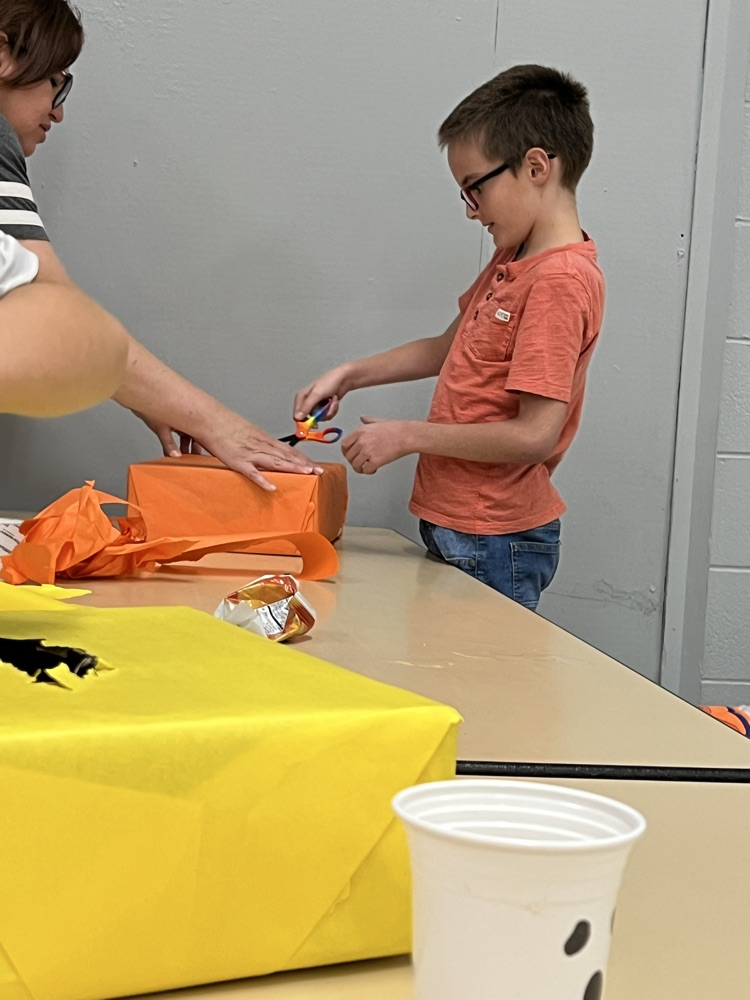 A boy with glasses cuts a bag with scissors while a person helps. A yellow bag and cup sit on a table.